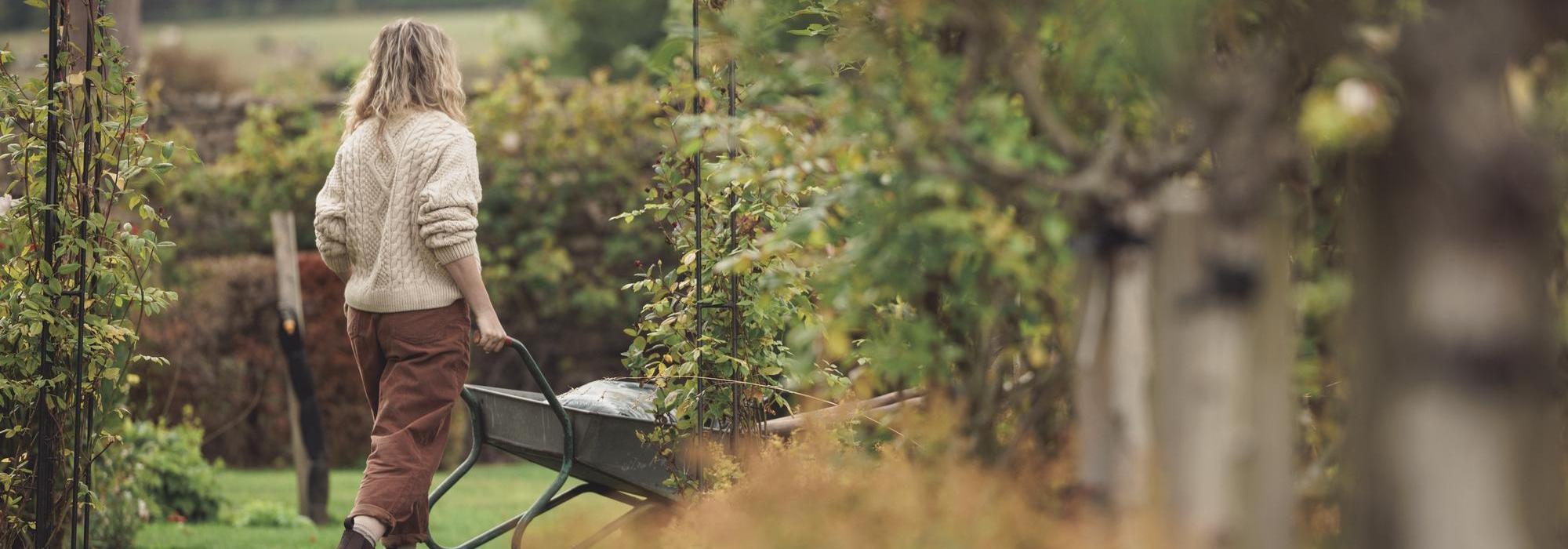Woman in a garden setting wheeling a wheelbarrow of gardening tools